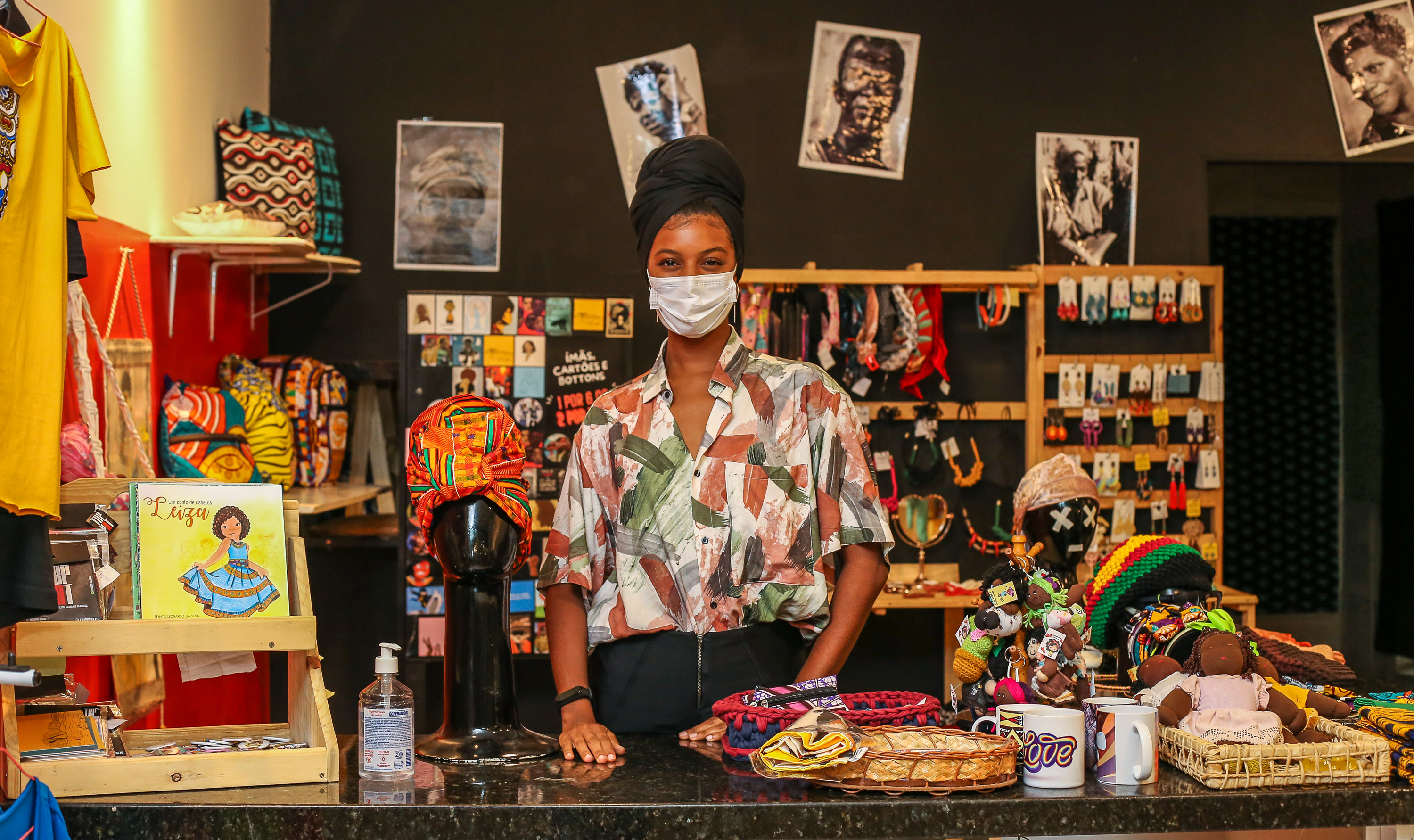 Mulher posa no estande da feira negra e podem ser vistos na foto:  artesanato, bijuterias, roupas, livros